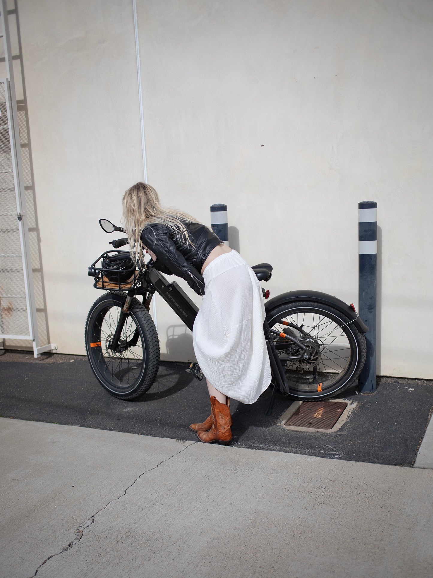 Person wearing a vintage leather jacket and cowboy boots who is bending over a parked electric bike in an outdoor setting.