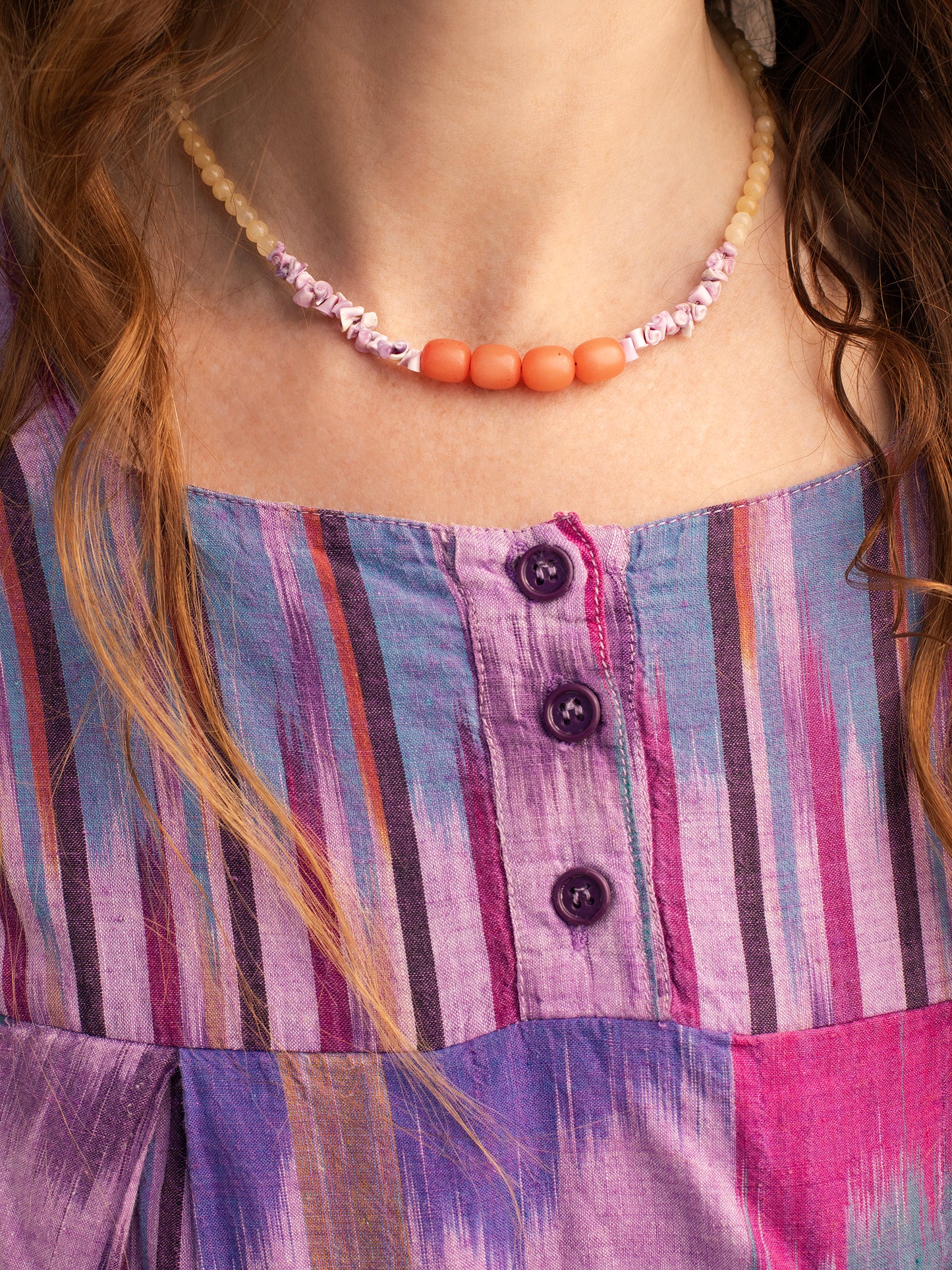 A necklace displayed on a person wearing a hand-woven purple & magenta ikat dress. The necklace features light yellow Citrine crystal beads, along with purple Sea Urchin shells & peach-colored Buri Seed beads.