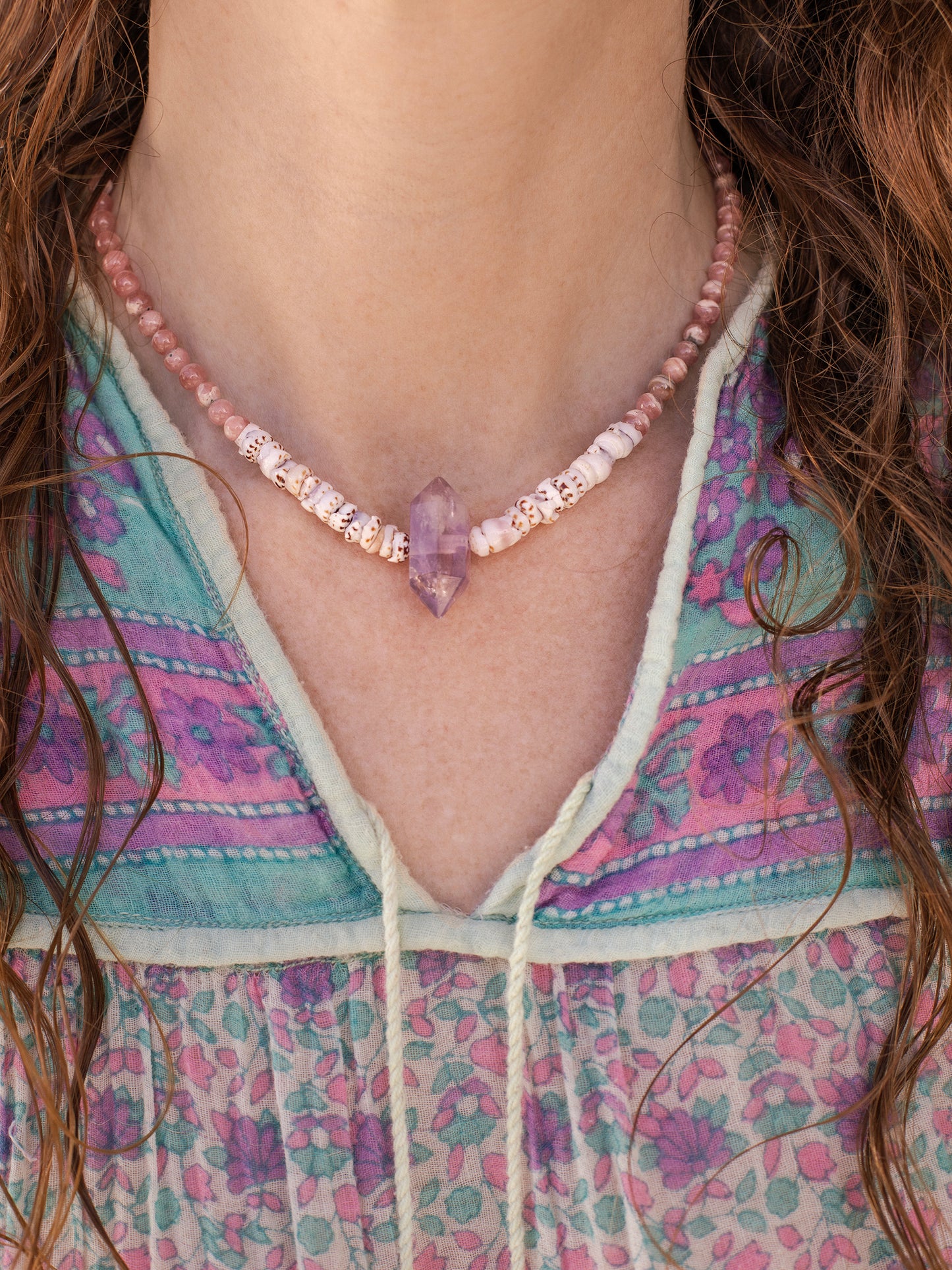 A necklace with a combination of pink Rhodochrosite, purple Amethyst stones, & natural Puka Shells, displayed on a woman wearing a vintage cotton gauze shirt & wavy hair.