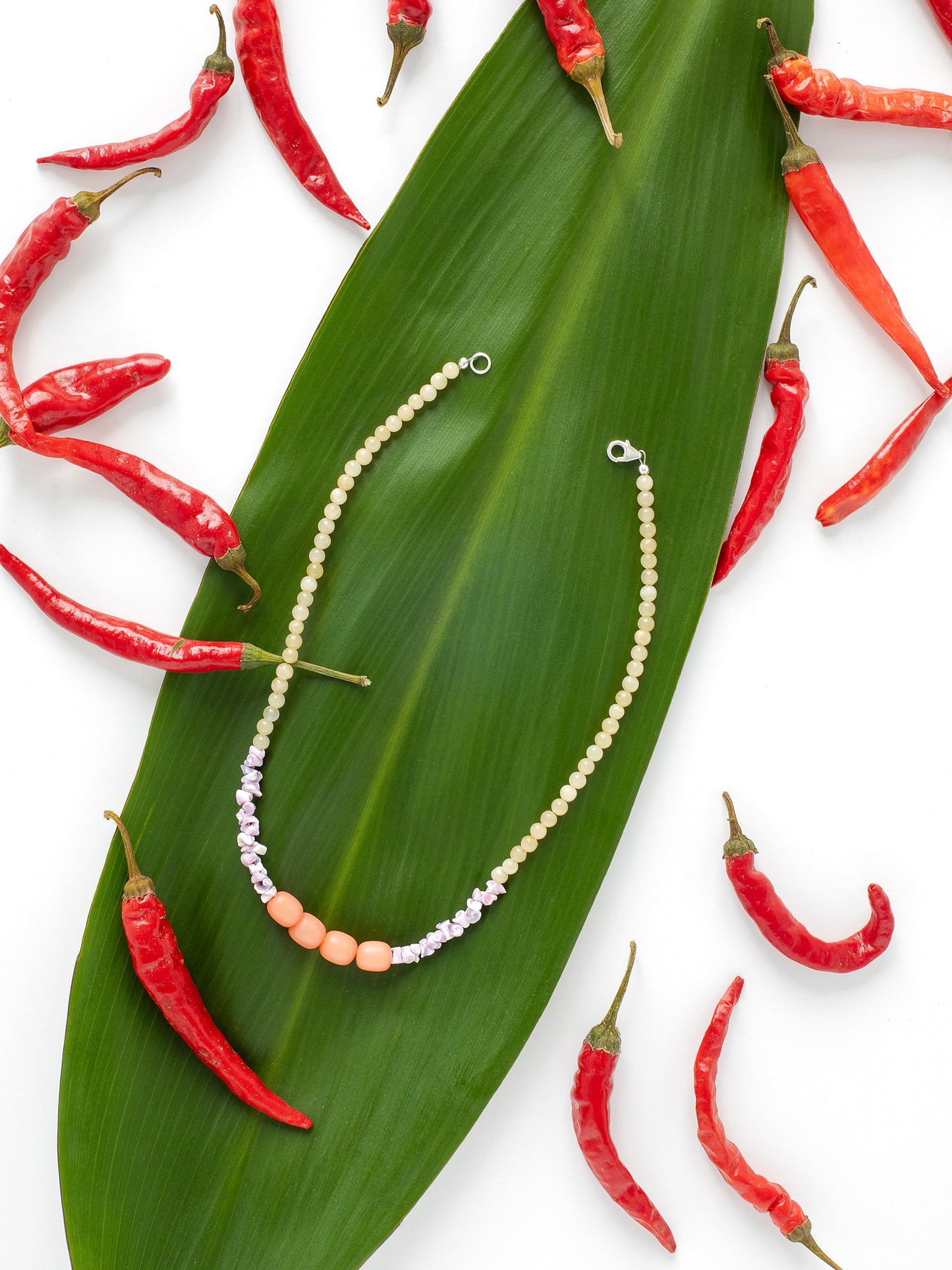 A necklace displayed on a green leaf with red chili peppers surrounding it. The necklace features light yellow Citrine crystal beads, along with purple Sea Urchin shells & peach-colored Buri Seed beads.