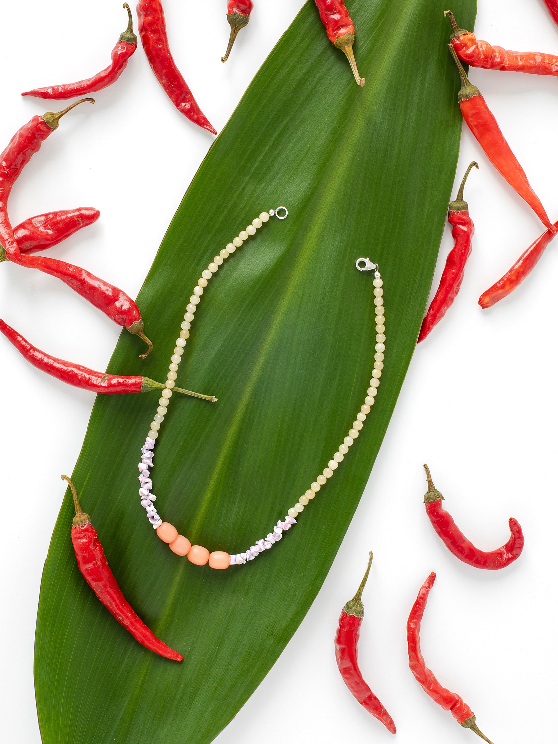 A necklace displayed on a green leaf with red chili peppers surrounding it. The necklace features light yellow Citrine crystal beads, along with purple Sea Urchin shells & peach-colored Buri Seed beads.