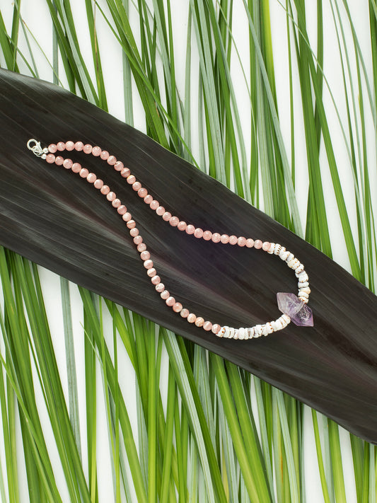 A necklace with a combination of pink Rhodochrosite, purple Amethyst stones, & natural Puka Shells, displayed on a leafy background.
