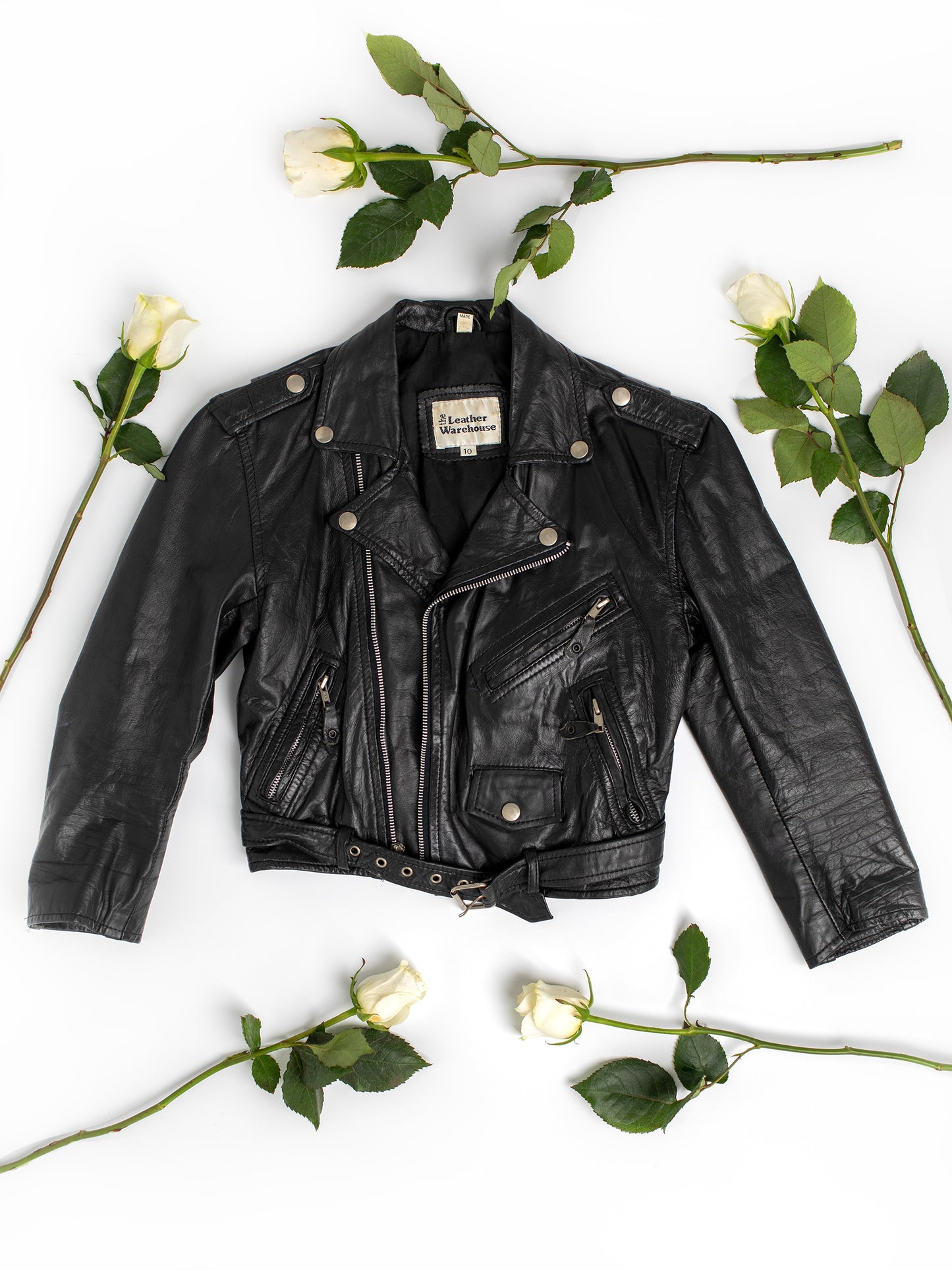 The front side of a vintage black leather motorcycle jacket on a white background with greenery and white long-stemmed roses.