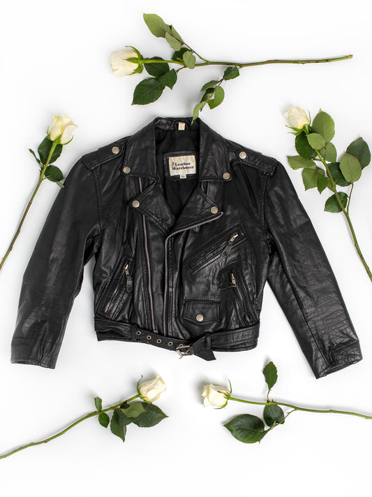 The front side of a vintage black leather motorcycle jacket on a white background with greenery and white long-stemmed roses.