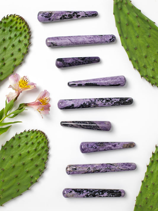 Nine polished purple Charoite crystal pointed massage wand amulets are arranged vertically on a white background, framed by green prickly pear cactus pads and pink flowers on the sides.