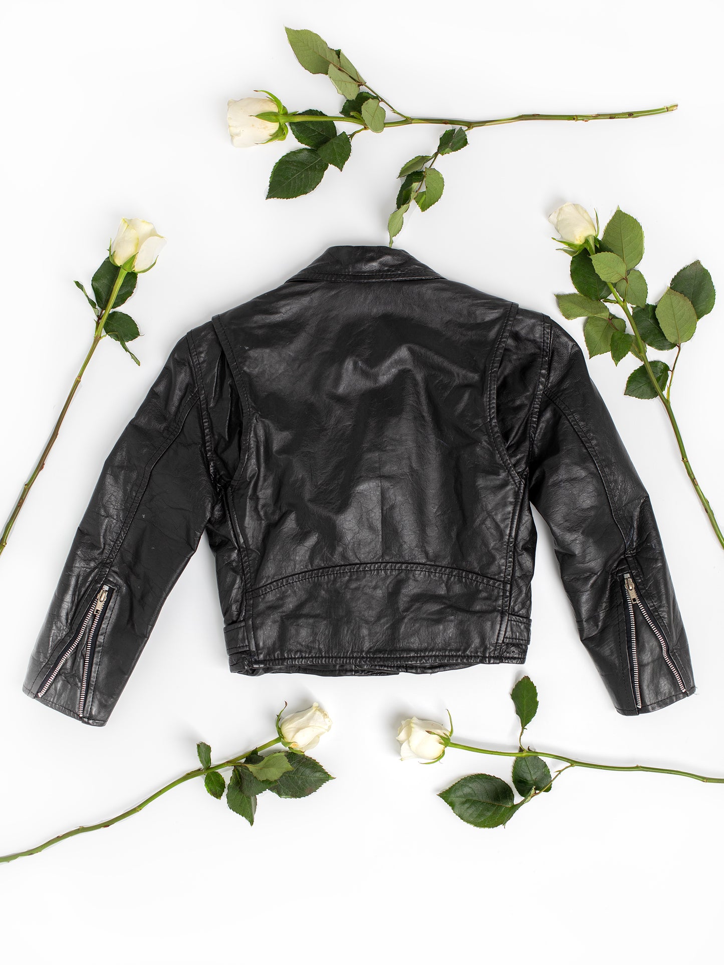 The backside of a vintage black leather motorcycle jacket on a white background with green leaves and white long-stemmed roses.