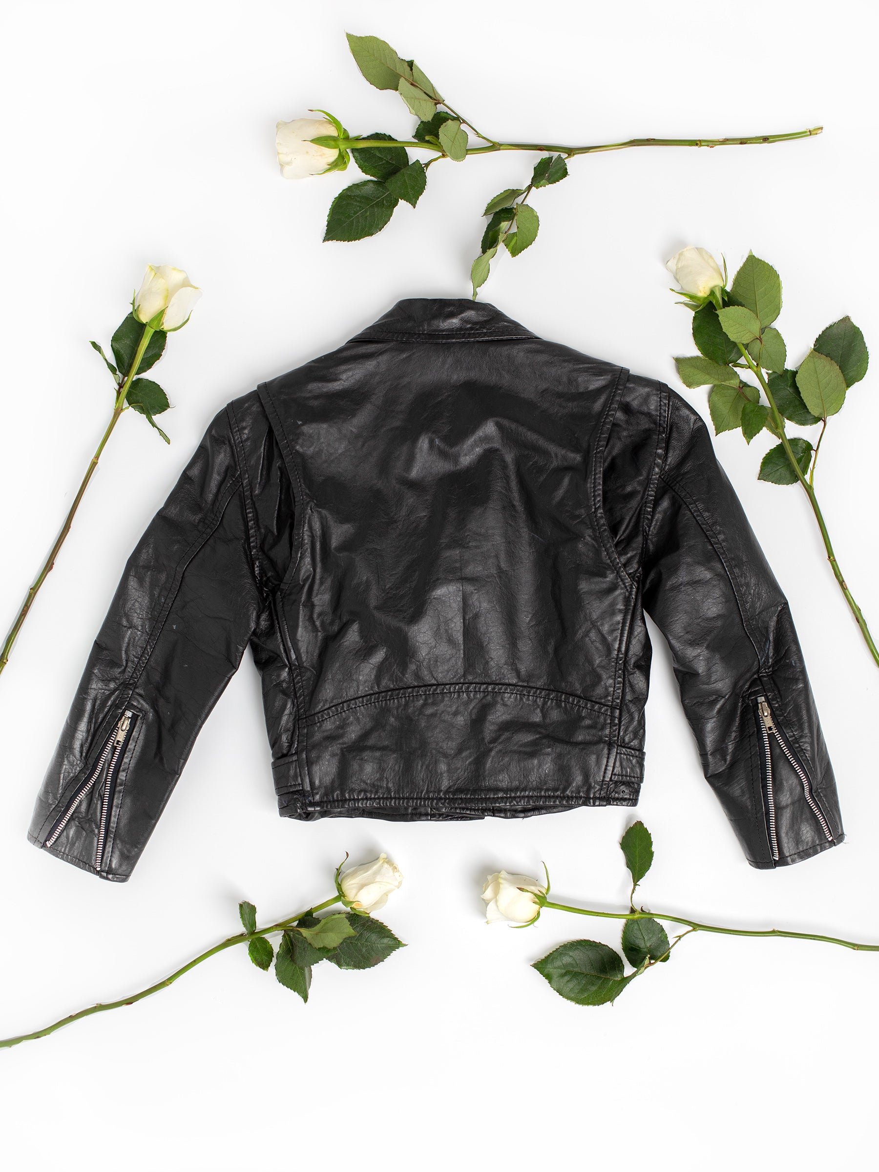 The backside of a vintage black leather motorcycle jacket on a white background with green leaves and white long-stemmed roses.