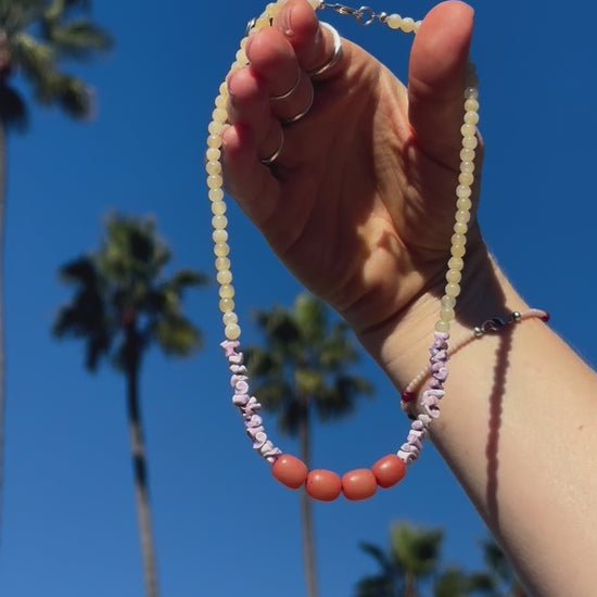 Video of delicate beaded necklace sparking in the sunshine amidst a backdrop of palm trees. The necklace features light yellow Citrine crystal beads, along with purple Sea Urchin shells & peach-colored Buri Seed beads.