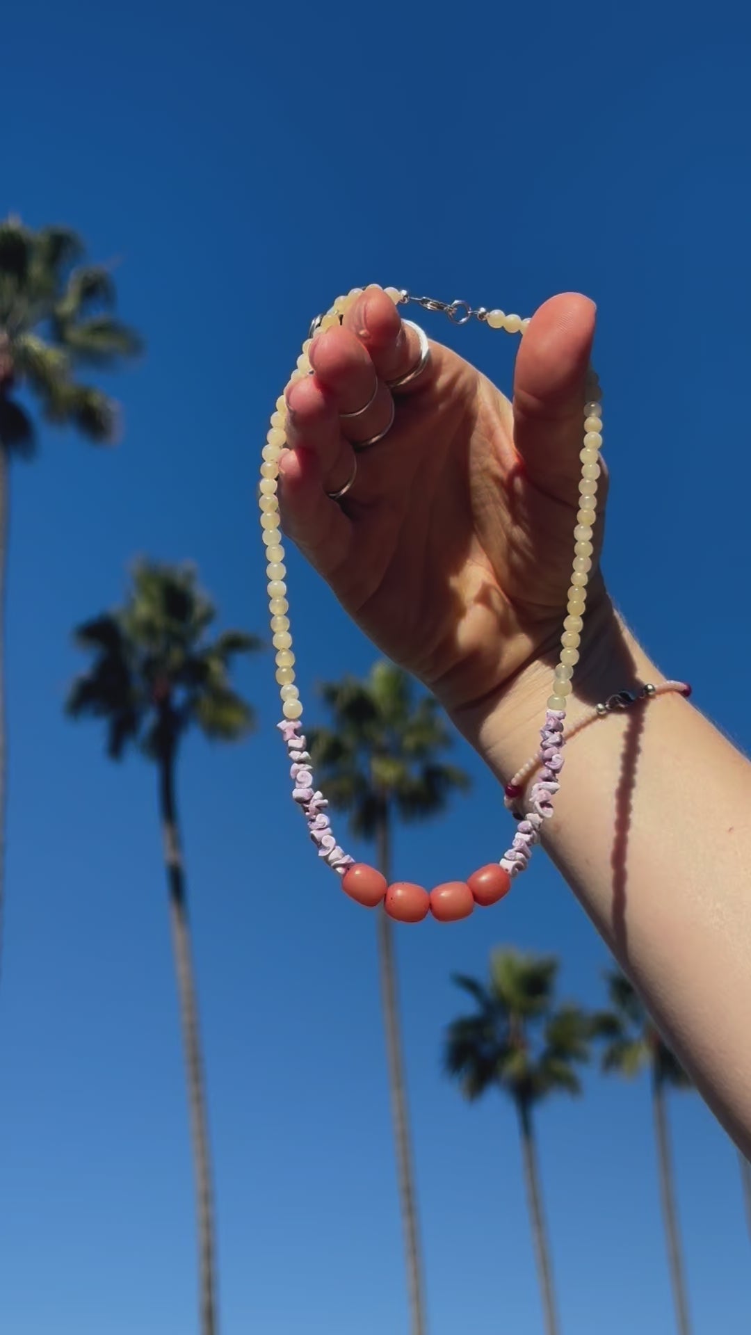 Video of delicate beaded necklace sparking in the sunshine amidst a backdrop of palm trees. The necklace features light yellow Citrine crystal beads, along with purple Sea Urchin shells & peach-colored Buri Seed beads.