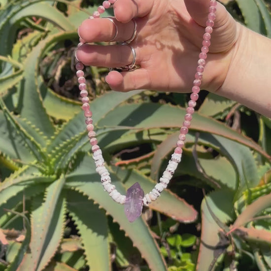 A necklace with a combination of pink Rhodochrosite, purple Amethyst stones, & natural Puka Shells, featuring a Sterling Silver clasp, against a backdrop of aloe vera succulents in the sunshine. 