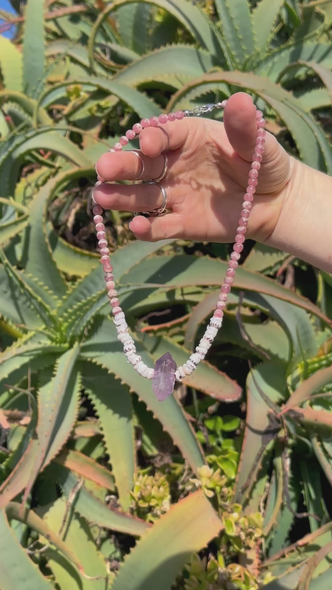 A necklace with a combination of pink Rhodochrosite, purple Amethyst stones, & natural Puka Shells, featuring a Sterling Silver clasp, against a backdrop of aloe vera succulents in the sunshine. 